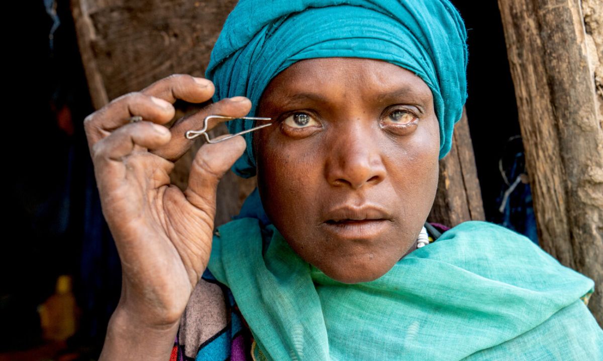 Eine Frau mit Kopftuch, die eine Pinzette nah an ihr Auge hält. 