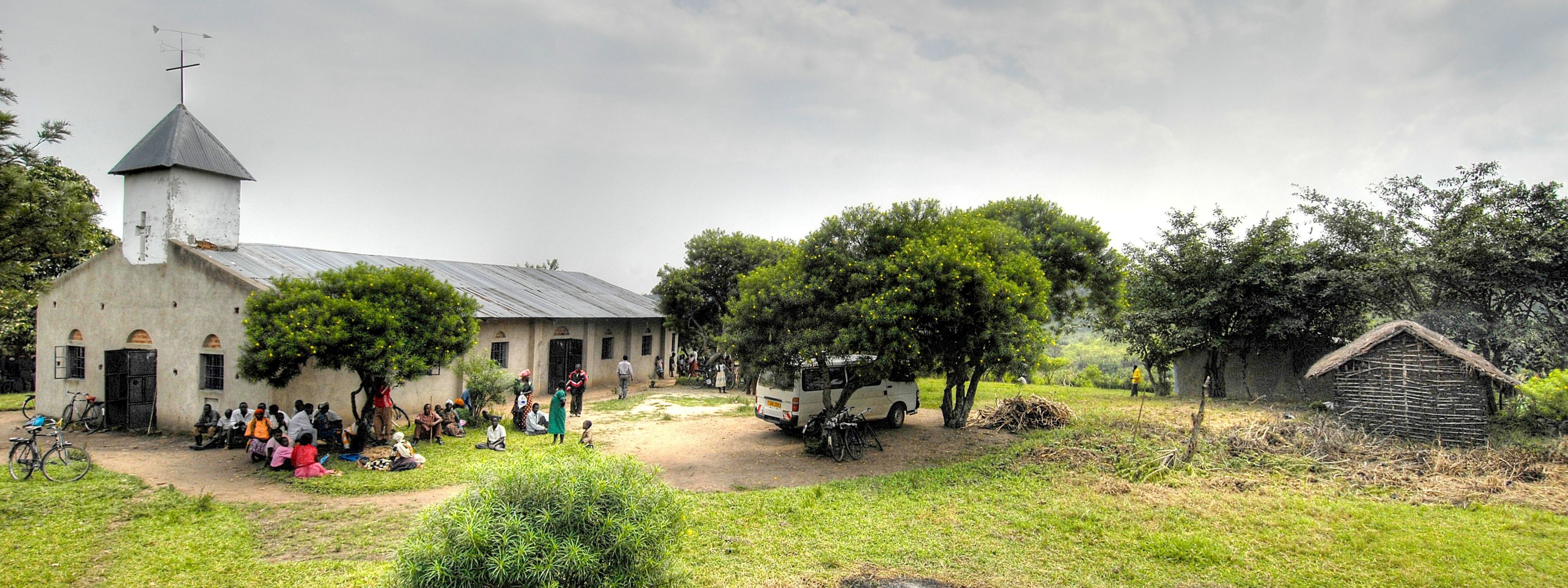 Landschaft mit Kirche, vor der eine Gruppe Menschen in bunter Kleidung sitzt.