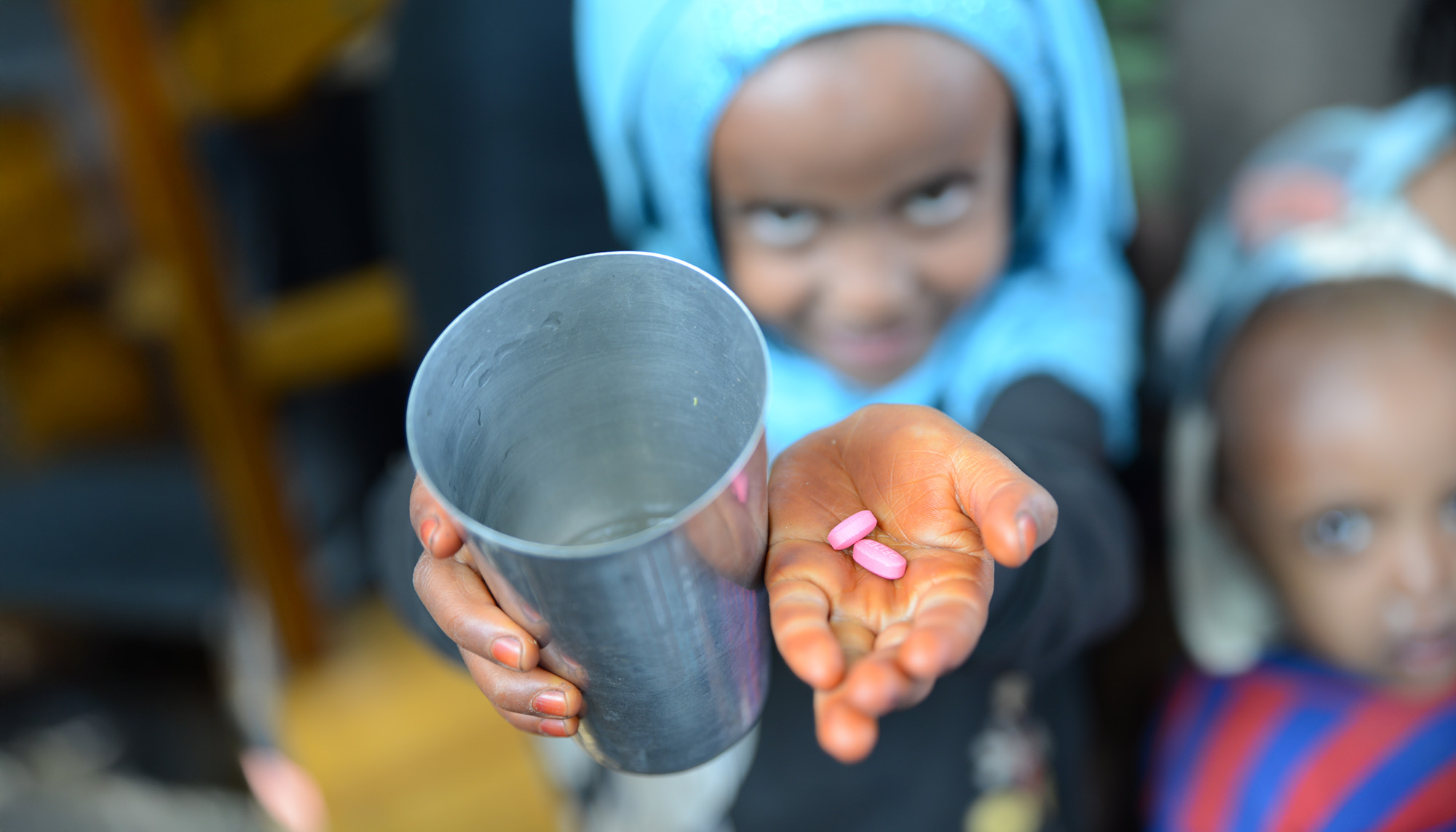 Ein Kind mit Kopftuch streckt die Hände hoch, in denen es einen Becher und zwei Tabletten hält.