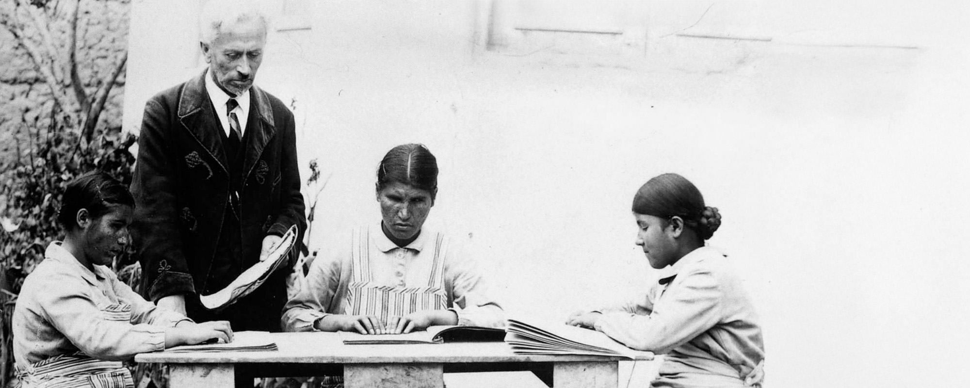 Drei Frauen sitzen an einem Tisch. Sie haben die Hände auf Schrifstücke gelegt. Ein Mann im Anzug steht daneben.