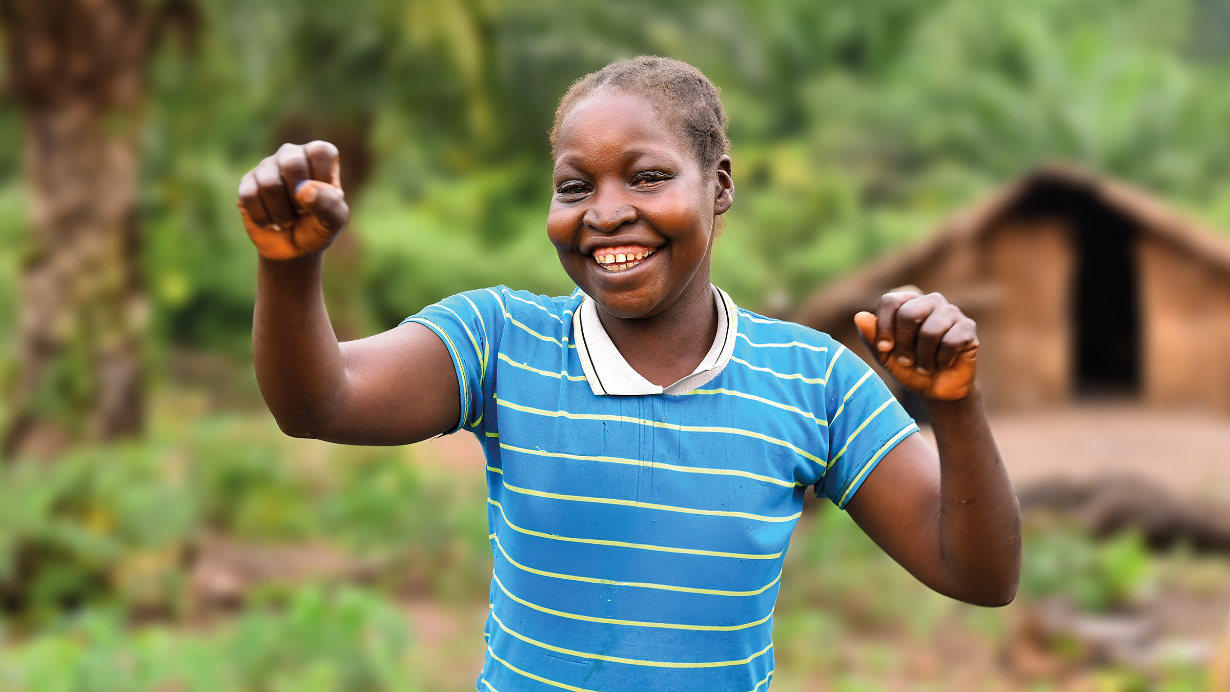 Eine junge, lachende Frau hält beide Hände hoch. Sie steht im Freien vor einer Hütte.